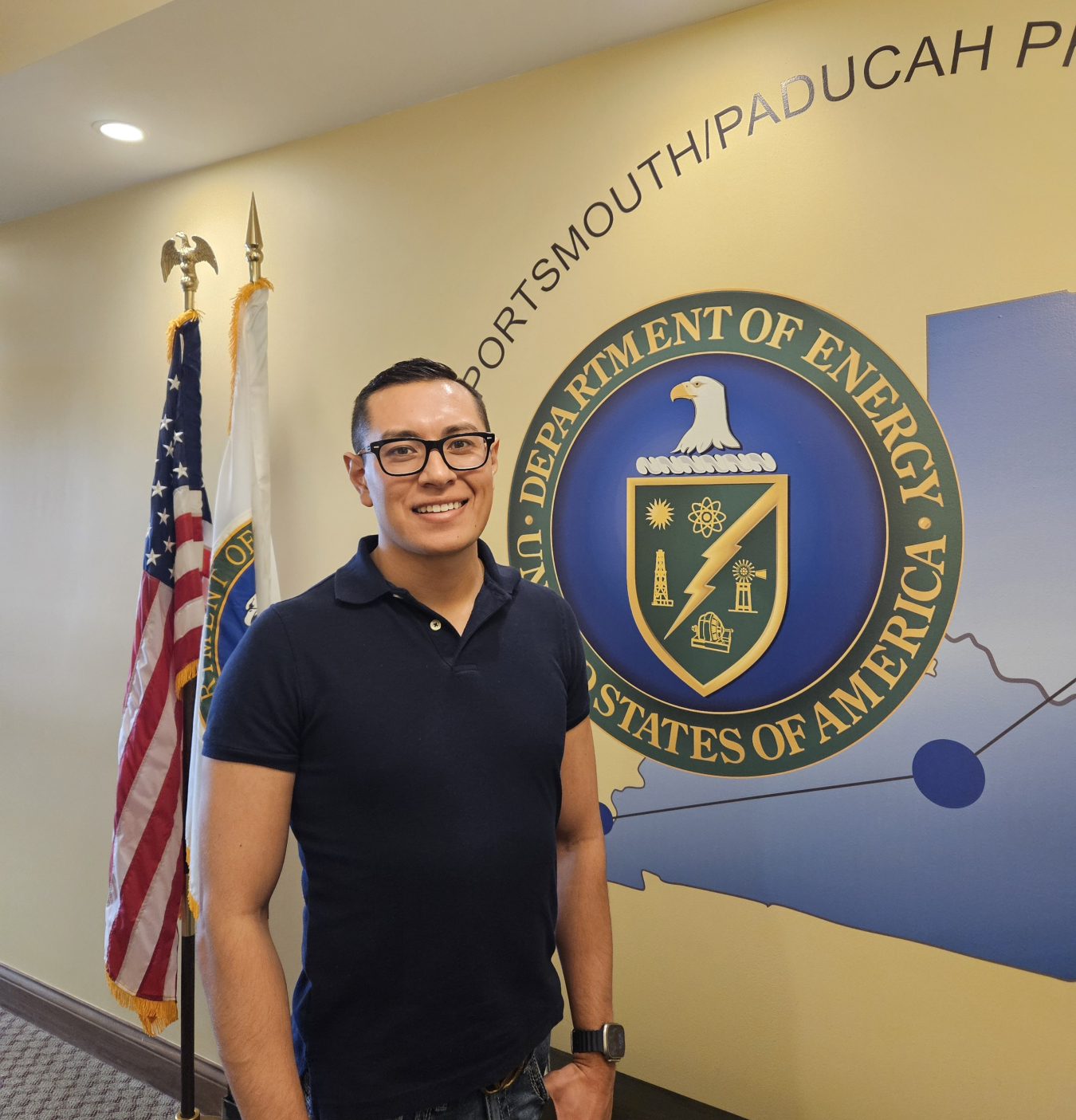 Man in black shirt stands in front of Department of Energy logo on wall.