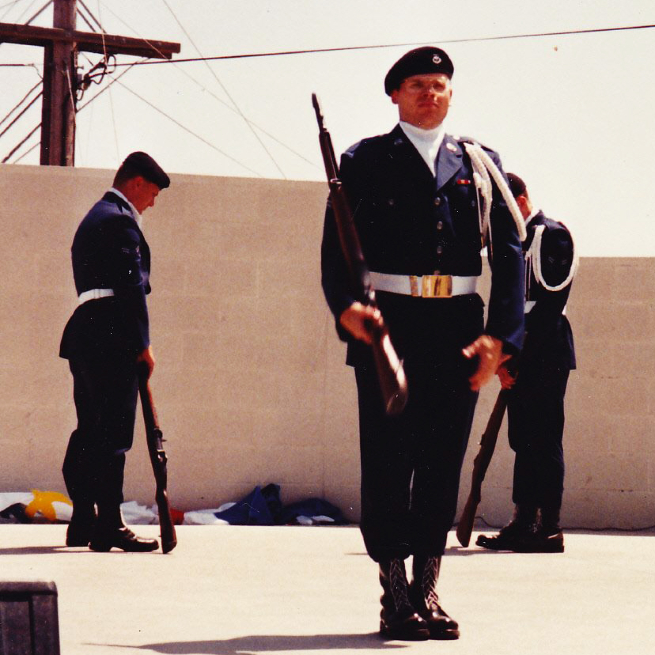 A man stands in a military uniform outside. Other people in uniform are nearby. He is at attention and is holding a rifle.