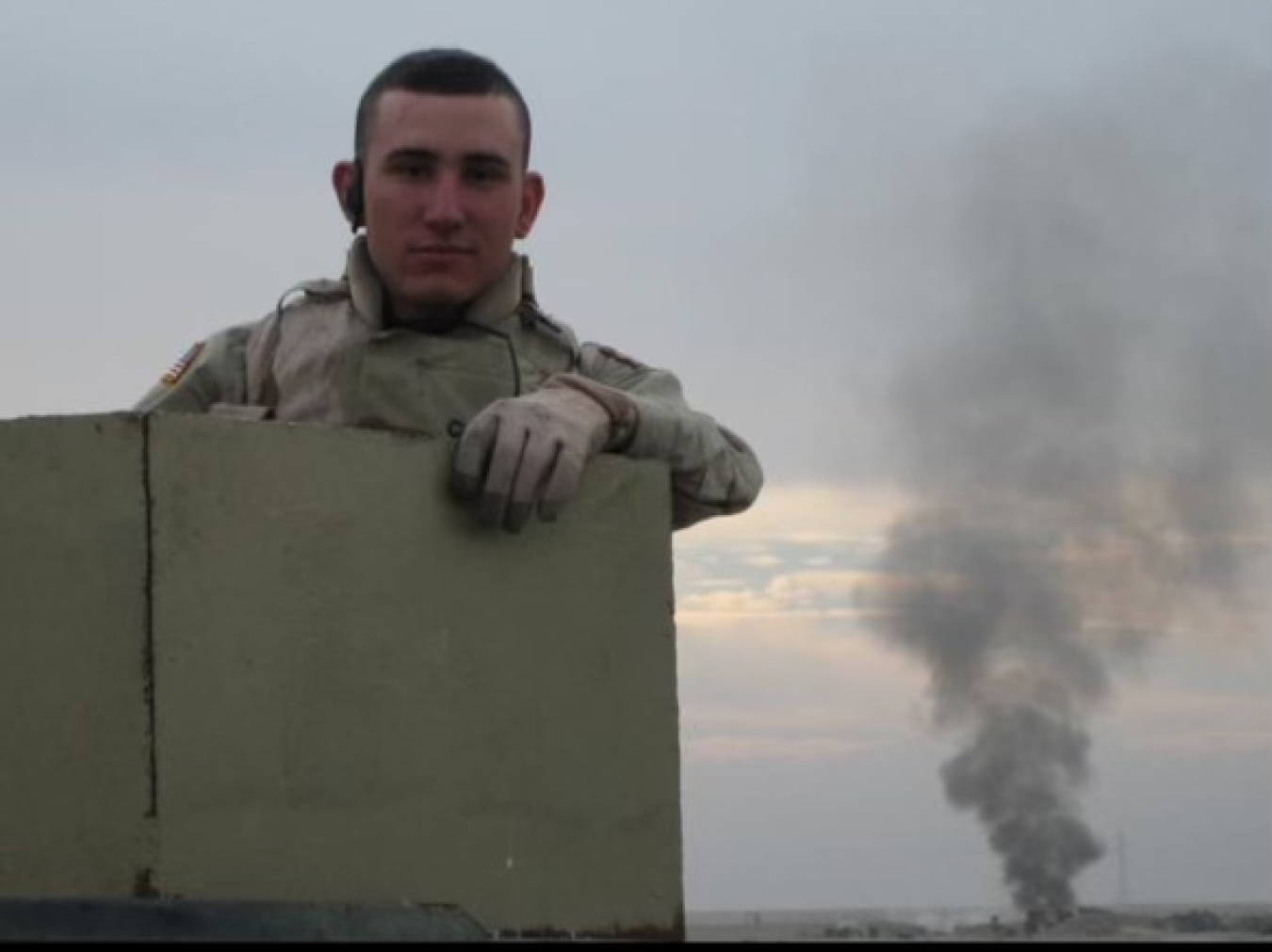 A man in a military uniform stands behind a short wall, a cloud of smoke can be seen behind him