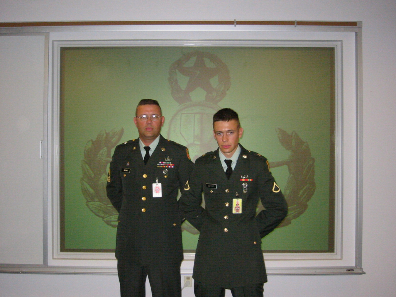 Two men in military uniforms stand and pose for a picture, they stand in front of a green and white wall with a black logo printed it