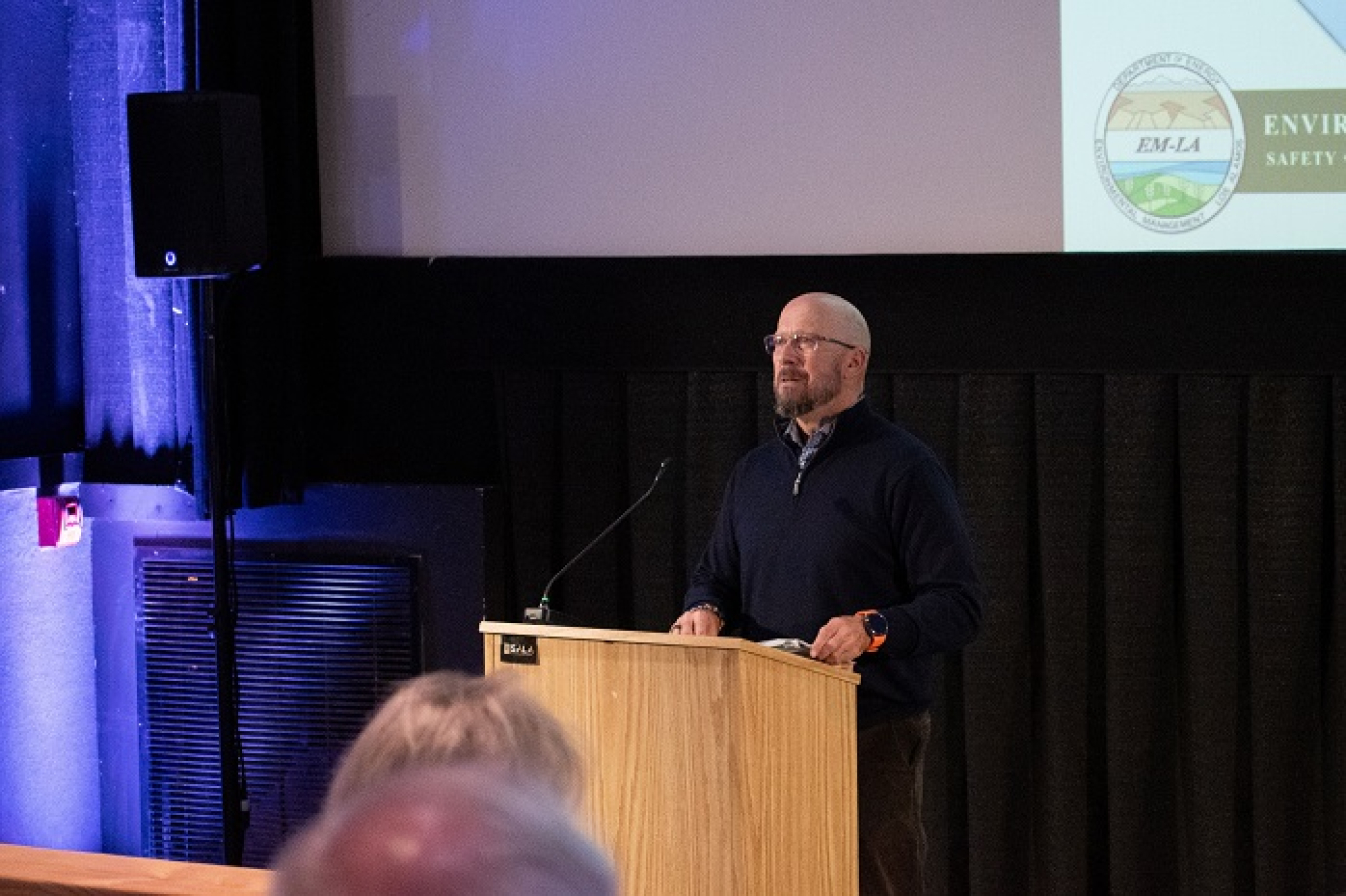 A man stands at a podium at the front of a room and addresses an audience
