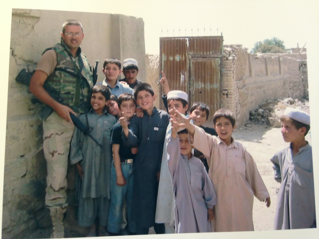 A man stands on the left against a wall with a camouflage military uniform, lots of children in gowns stand beside him and smile