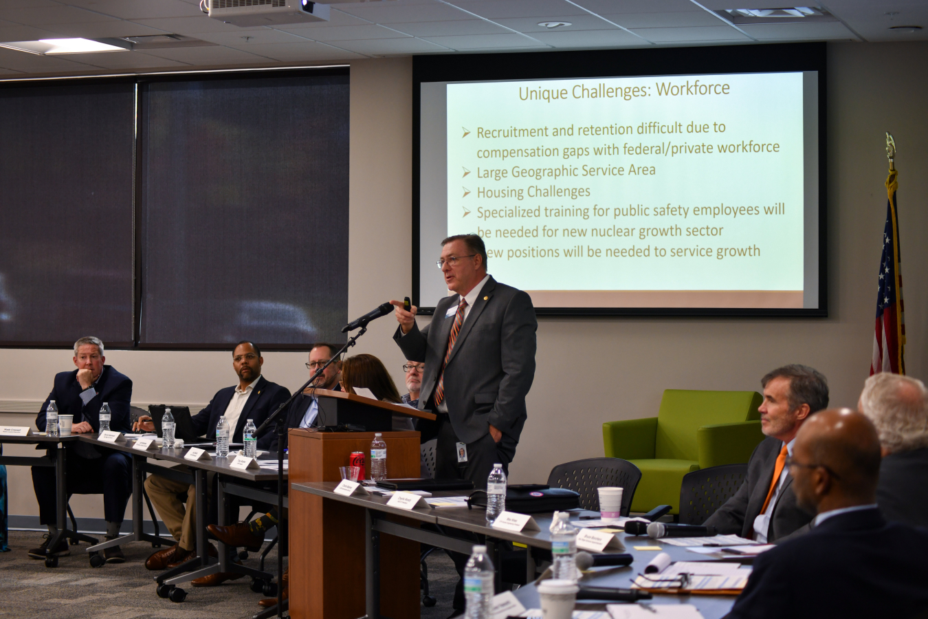 A panel of professionals sitting at the front of a room speaking to a crowd, one man stands at a podium in the middle of the front of the room