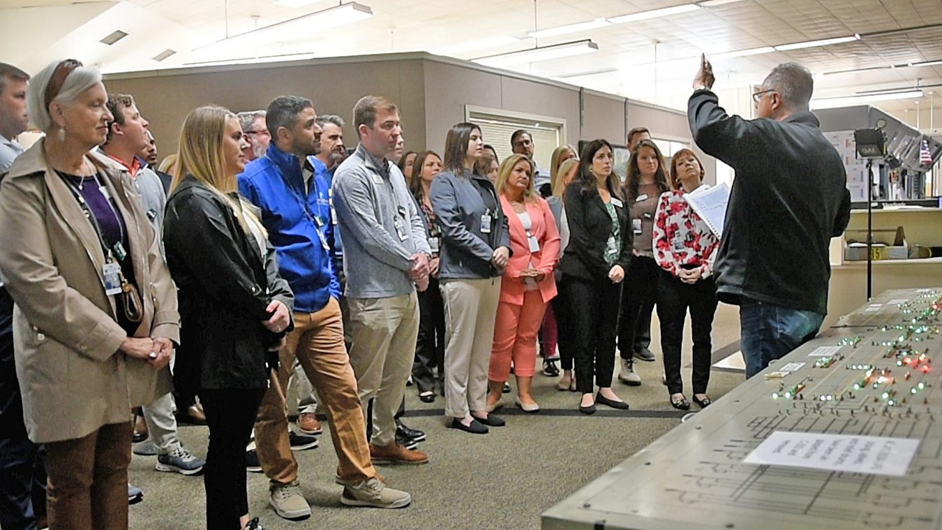 A group of adults stand in a room and listen to a man at the front of the room speak while giving a tour