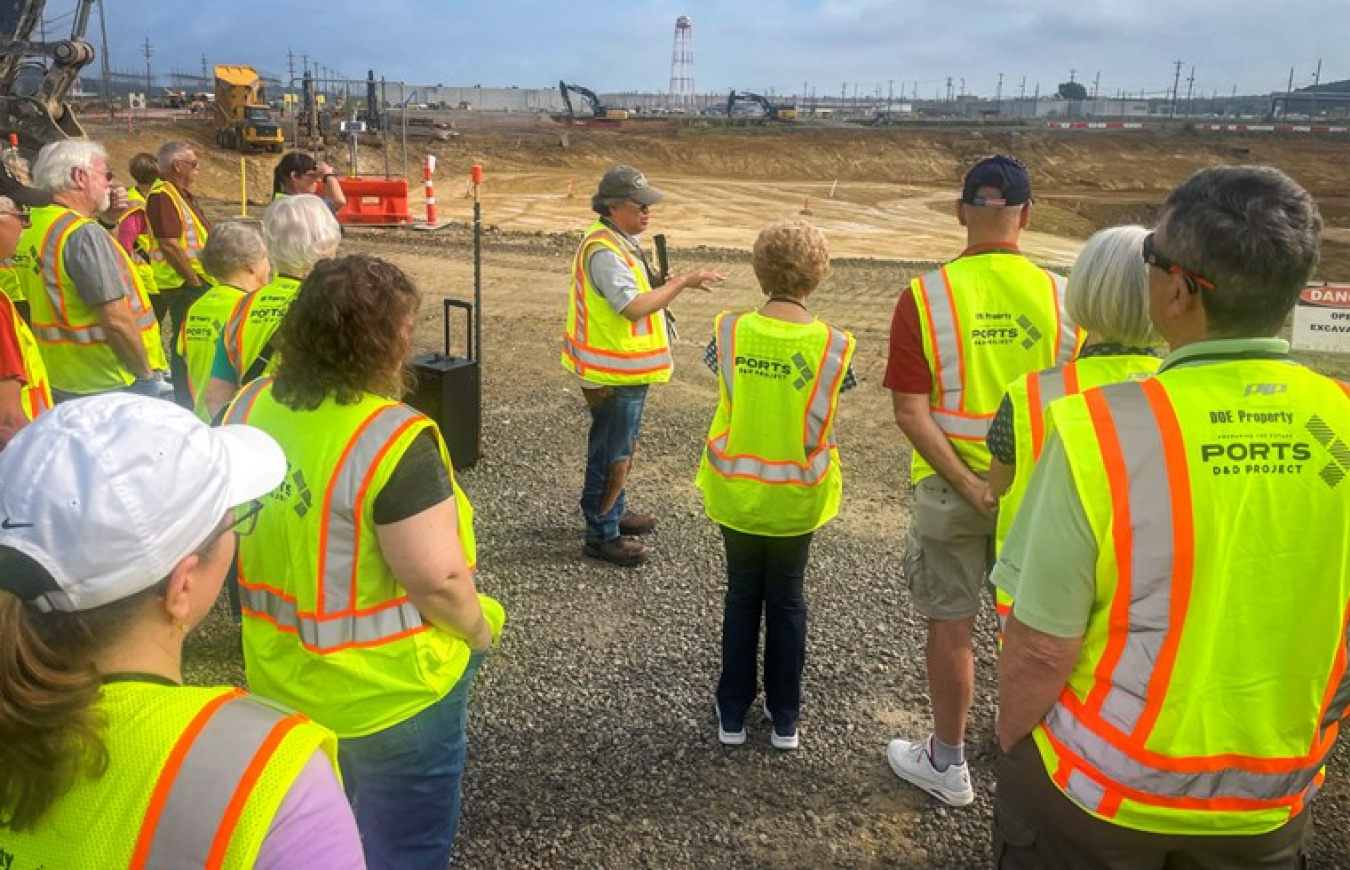 A group of people in bright yellow protection gear stands around outside and listens to a man leading a tour