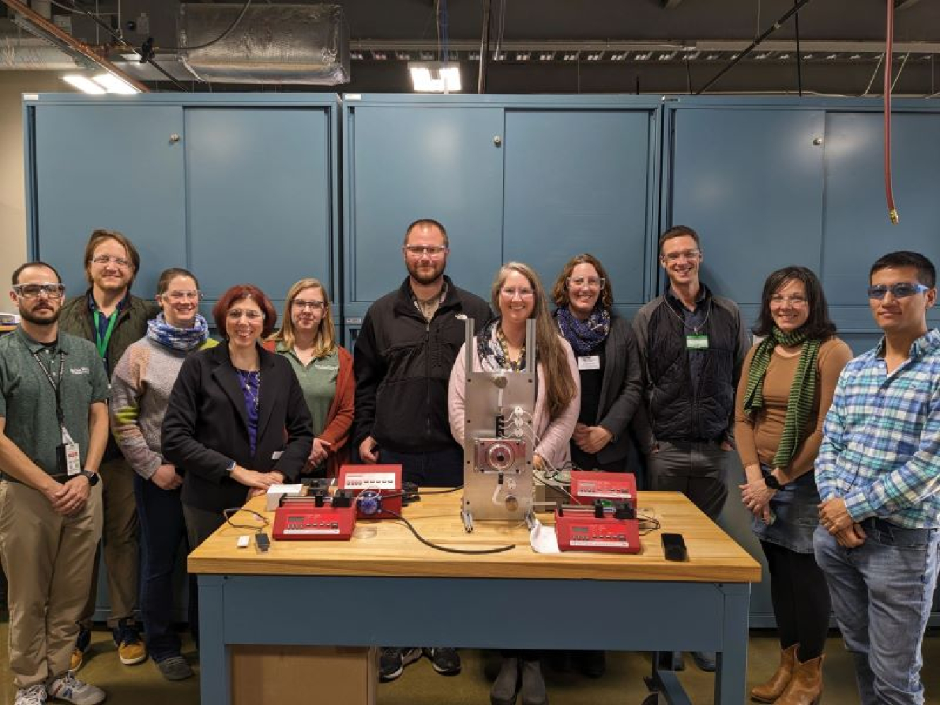 Moody and 10 other researchers standing behind a table that holds a metal contraption the size of a coffee machine.