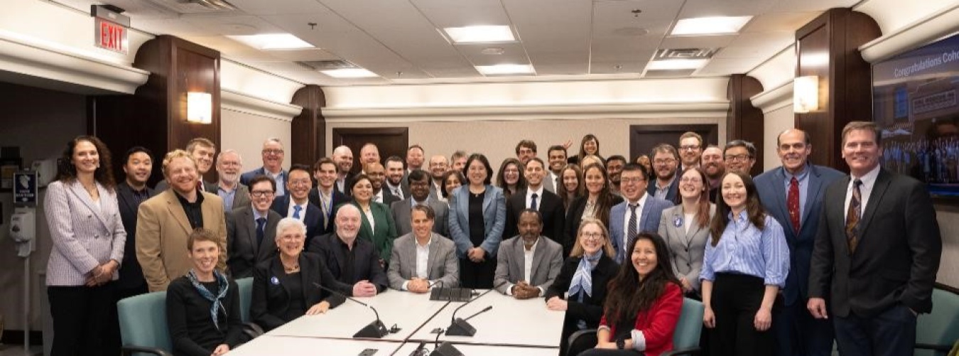 A group of 46 people in business attire pose and smile for the camera in a meeting room.