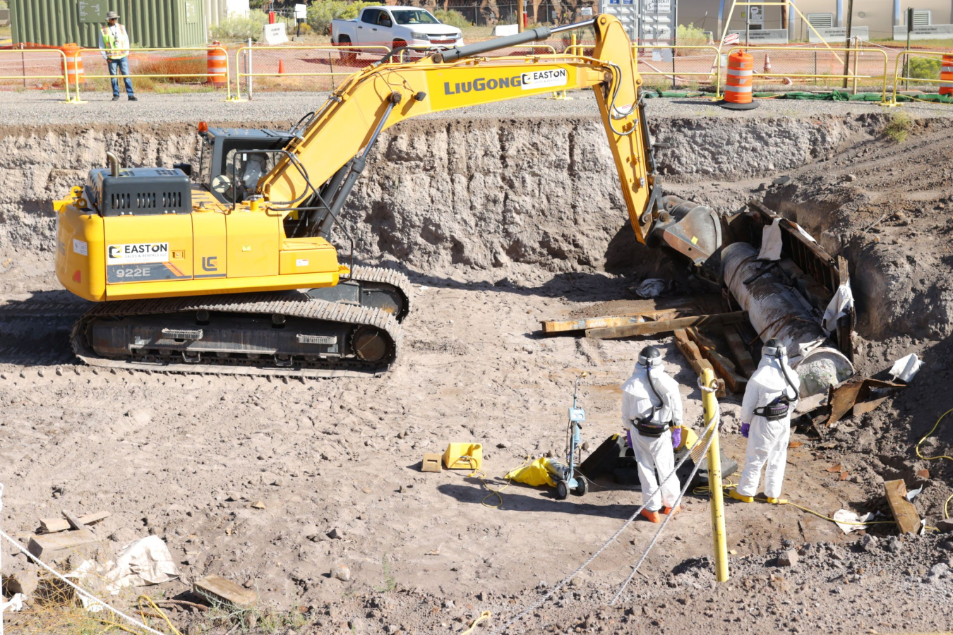 People in hazmat suits with excavator retrieving cemented container.