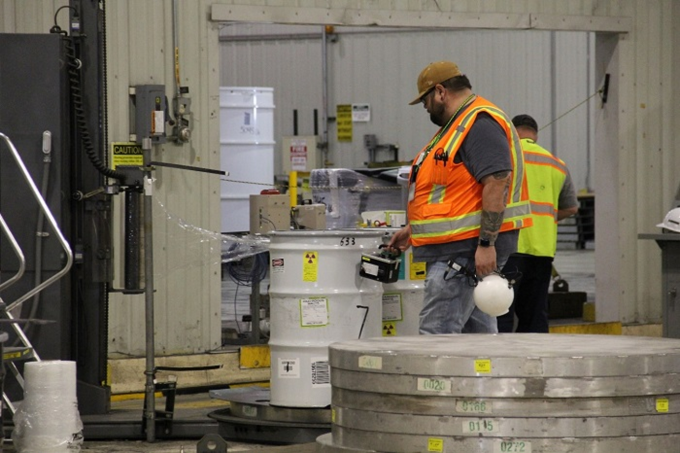Employees in protective gear work in a warehouse and inspect storage drums