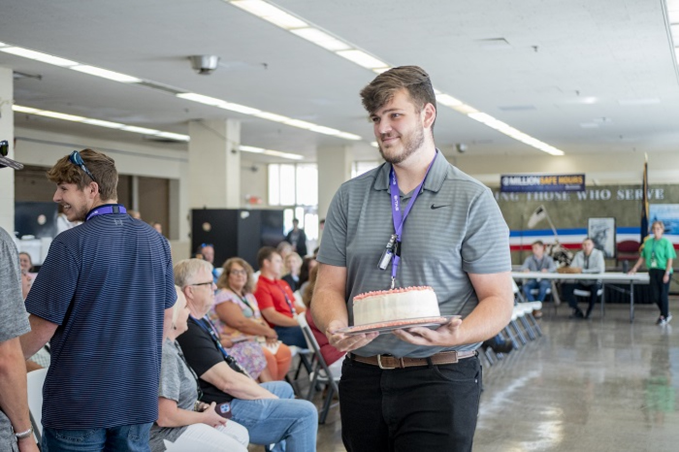 A man in a grey shirt holding a white and pink, round cake walks through a room 