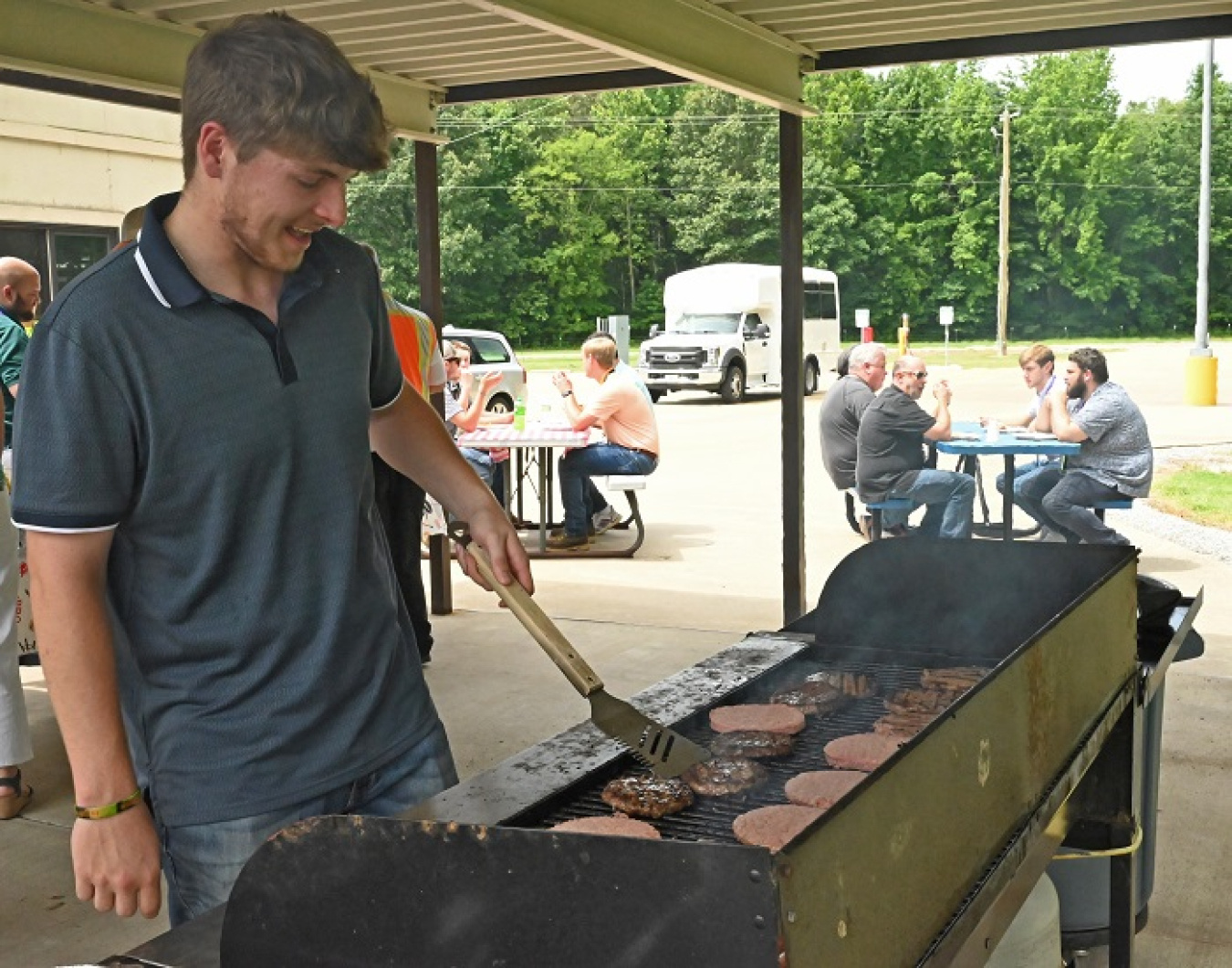 A man in a grey shirt stands in front of a grill and flips hamburgers with a spatula