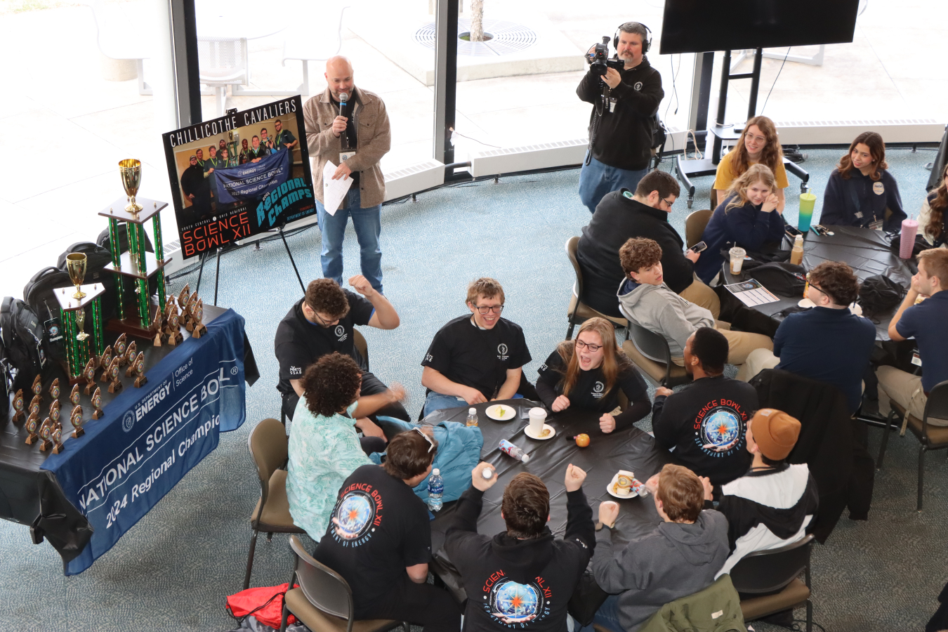 An overhead shot of tables with students sitting at them and a table filled with trophies
