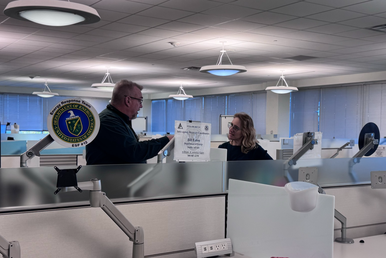 Two employees talking to each other inside an office building, the DOE logo is seen off to the left