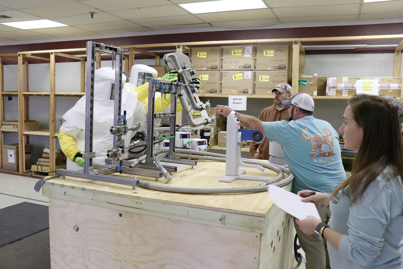 Employees in white hazmat suit perform procedures on piping, other employees stand around and observe