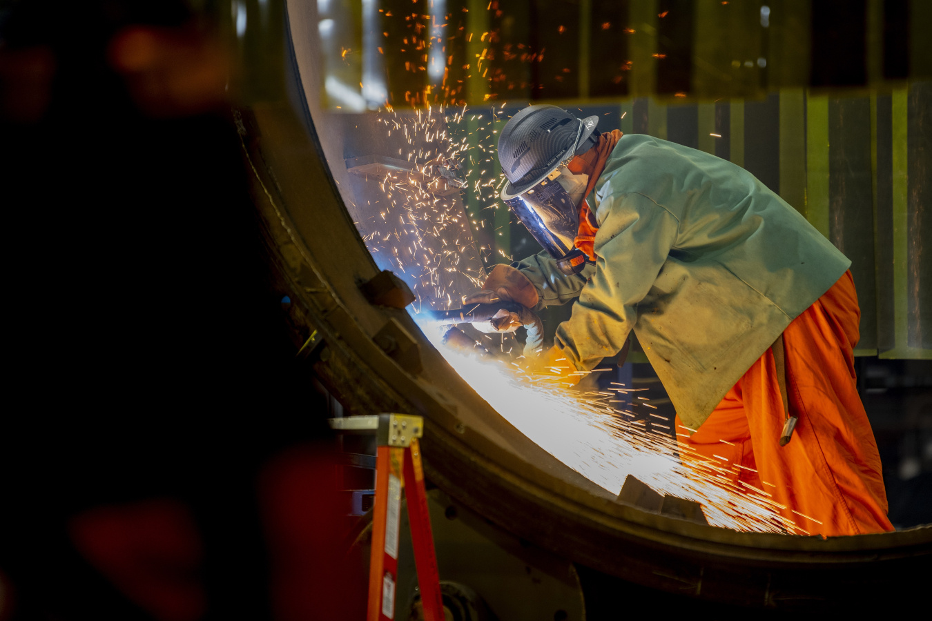 An employee wearing safety attire uses a welding machine inside a building, sparks fly off the object around him