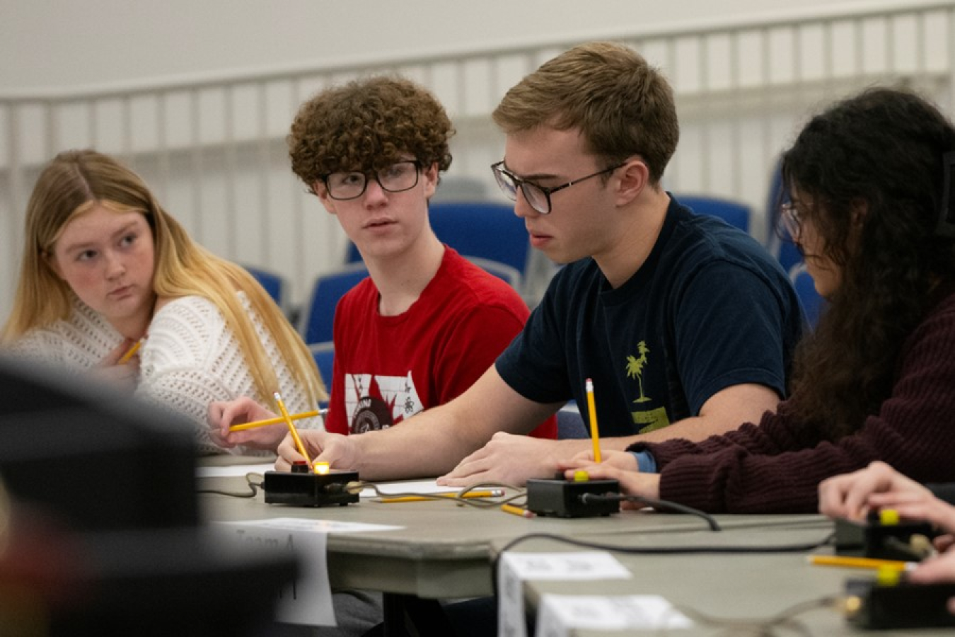 A group of students sit at a table and hold pencils while looking at each other