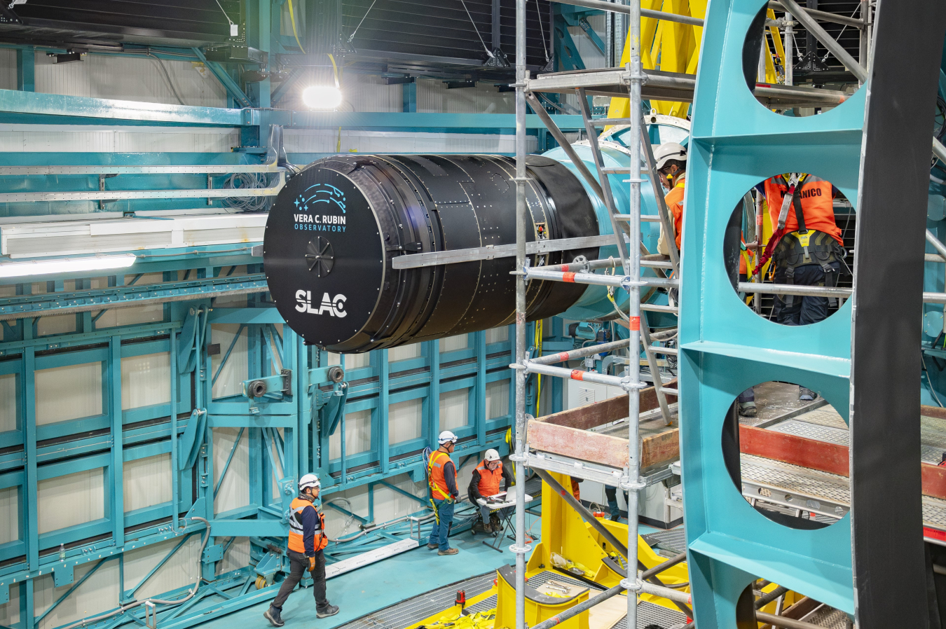 The giant telephoto lens of the LSST Camera as it is being placed on the Rubin Observatory, surrounded by scaffolding. People are below it in orange safety gear.