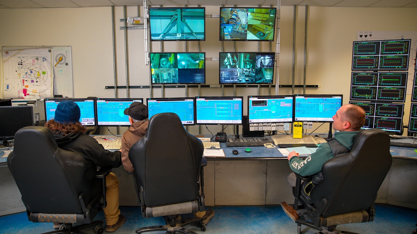 A group of people sitting in chairs at a control room desk that holds lots of computers and screens