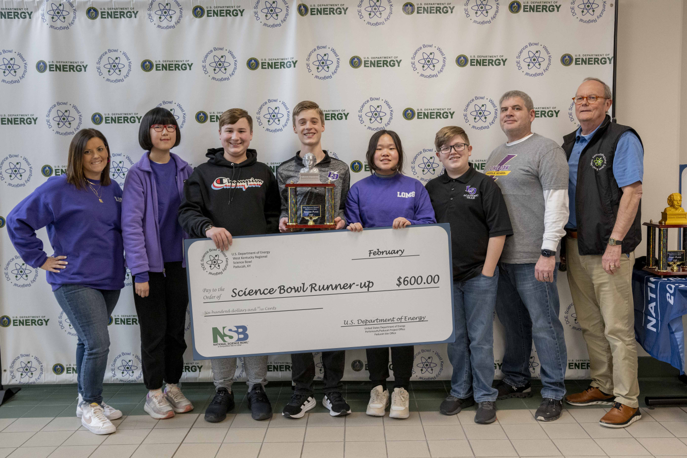 A group of midde school students holding a banner and trophy's after winning a science bowl competition