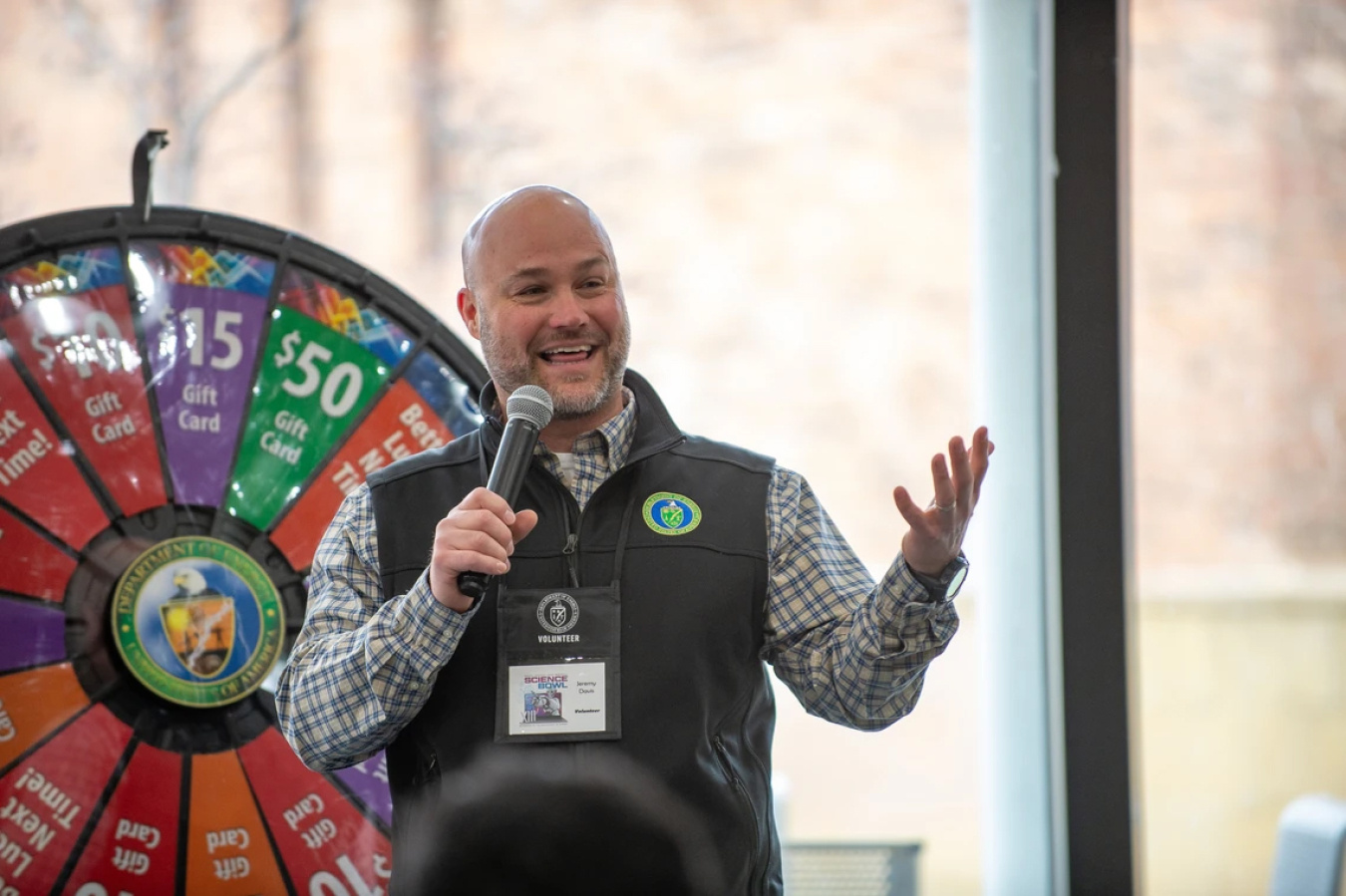 A man with a microphone talks to a crowd with a spin-to-win wheel behind him