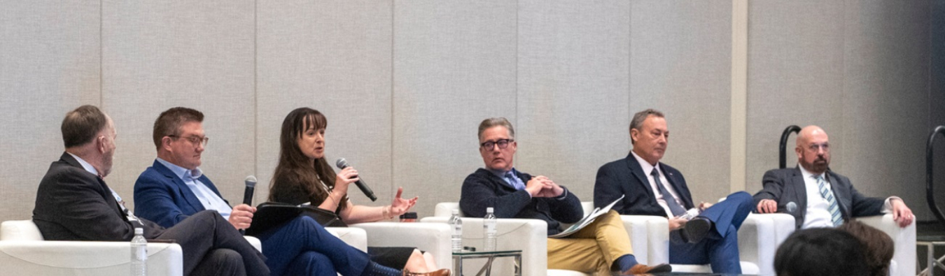 A group of six people sitting in white chairs on a stage for a panel at a conference