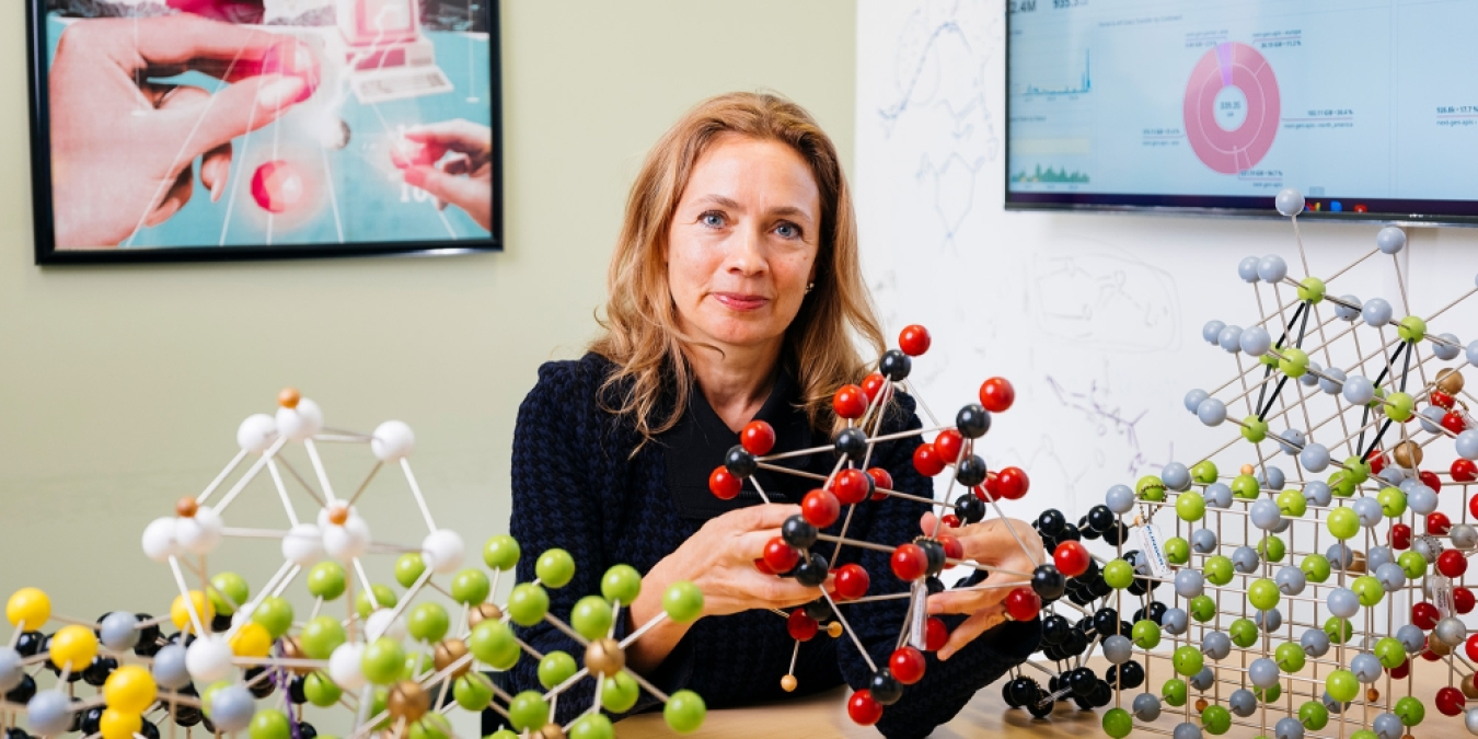 Kristin Persson (a woman in a black shirt) sitting at a desk in an office. She is holding a complex molecular structure and there are several molecular structures on the table in front of her. 
