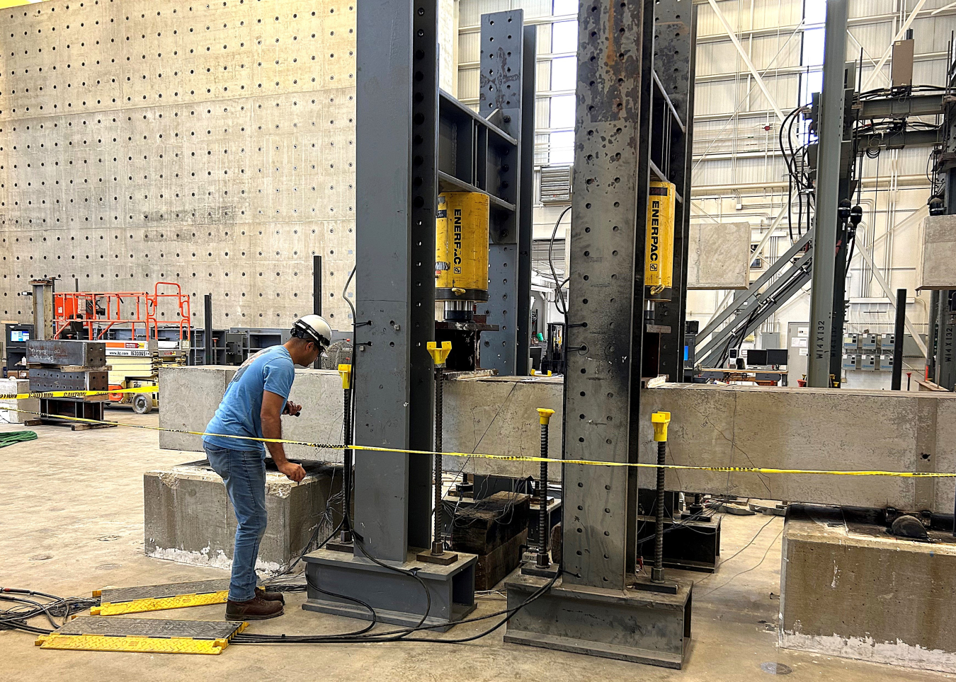 man in blue shirt and white helmet stands next to horizontal column of gray concrete inside test facility