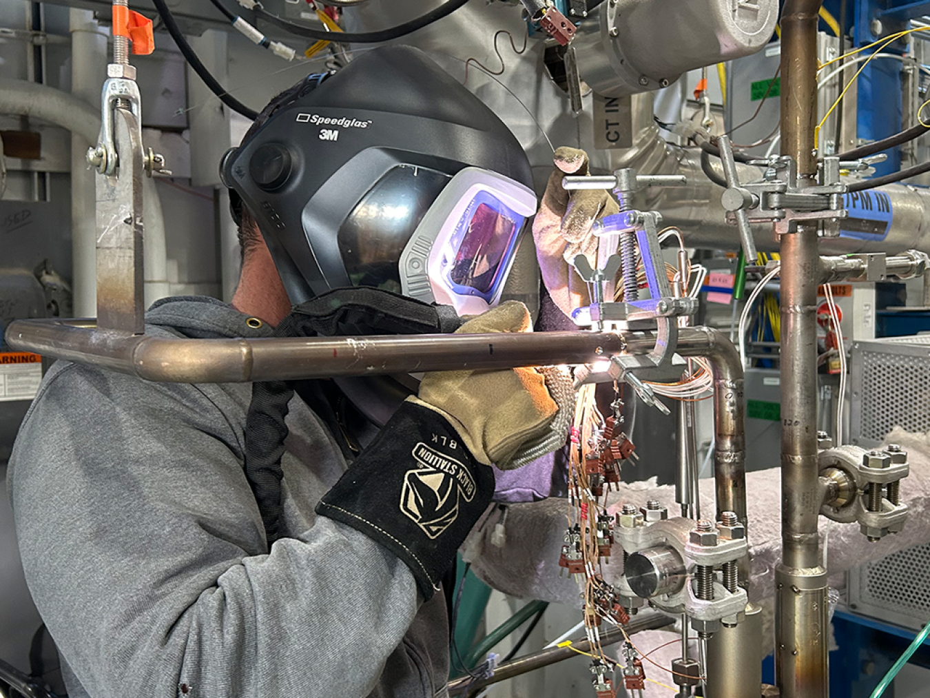 A worker wearing a welding mask welds a pipe onto the new cold trap device at the METL facility.