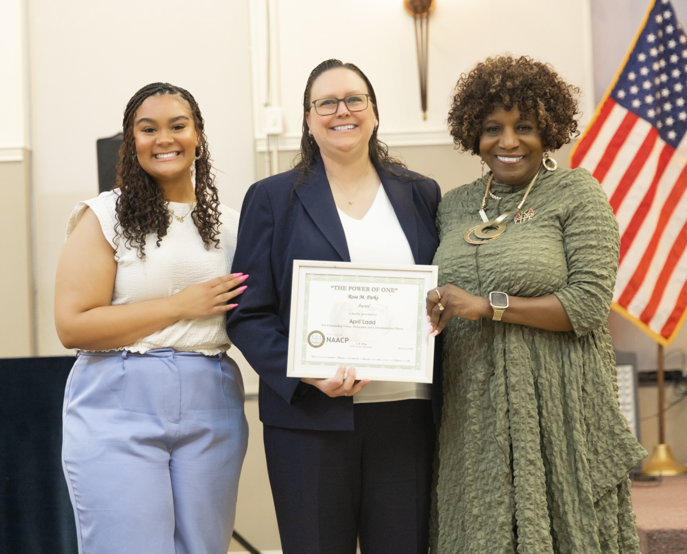 Three women smile for a group picture, one holds up an award certificate