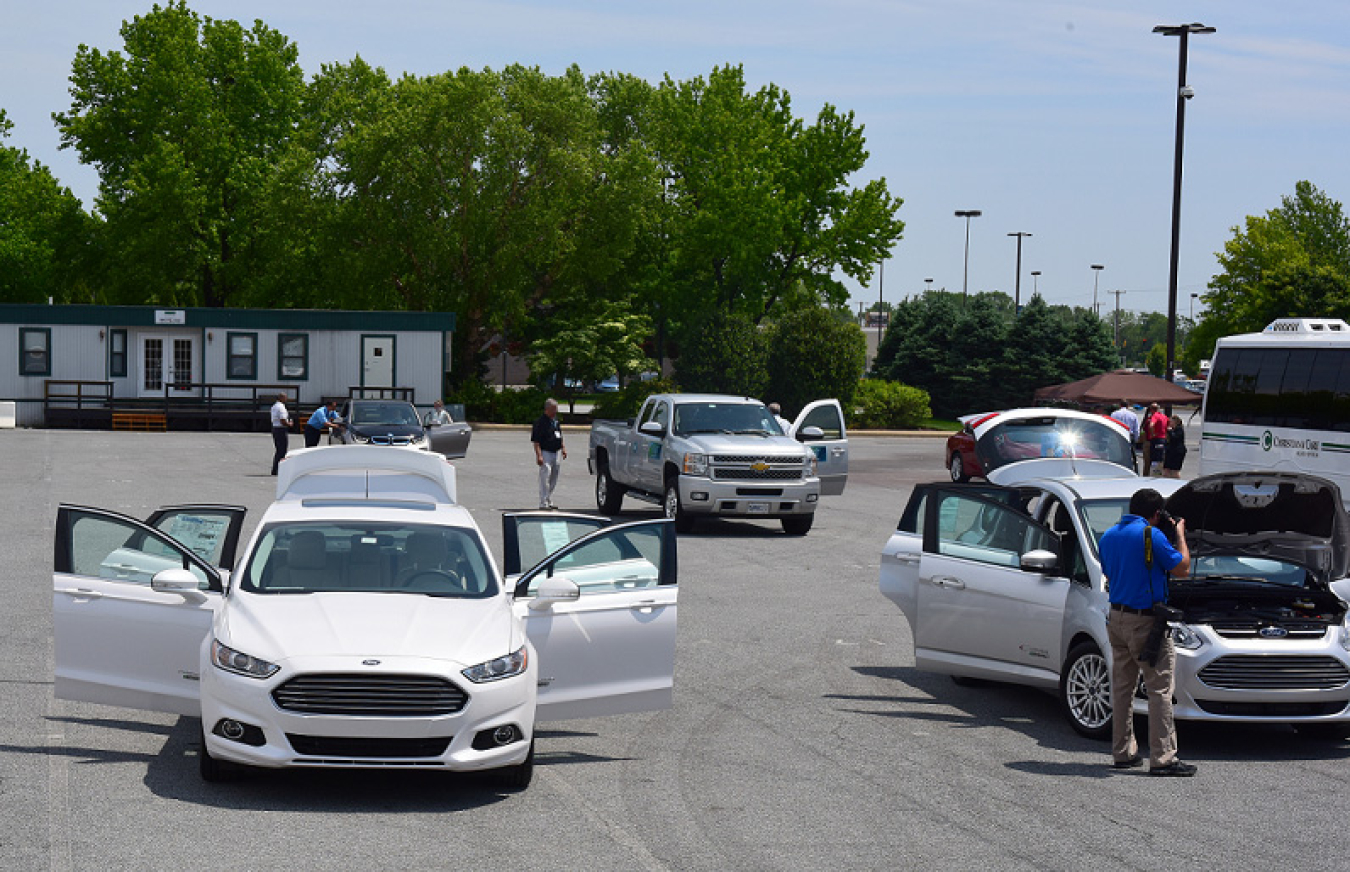 A variety of alternative fuel vehicles in a parking lot with several people looking at them.
