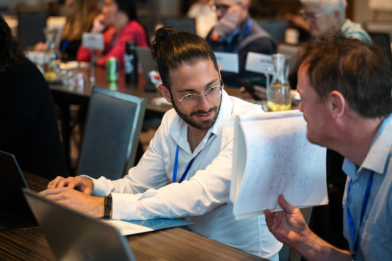 Two men are engaged in conversation, while one holds a notebook up for the other to see.