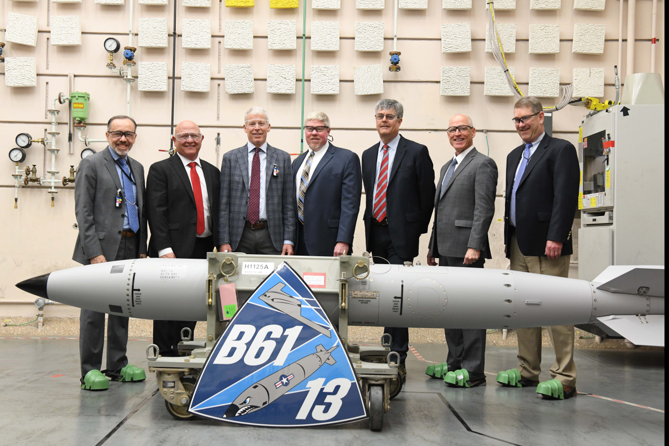 A group of men stand behind the first B61-13. In front of the bomb is an enlarged print of the B61-13 logo