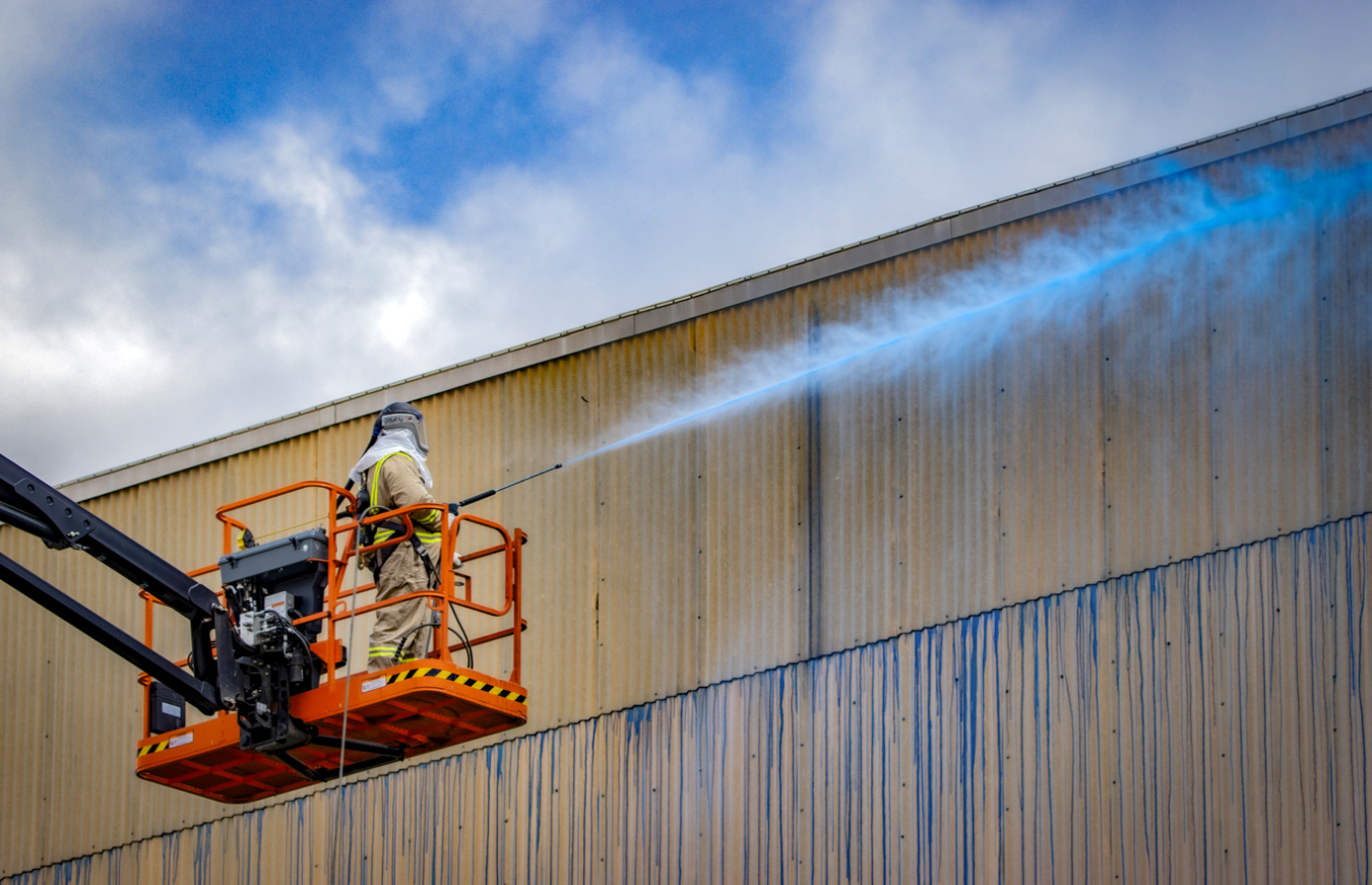 A worker on a lifted work machine sprays a blue substance onto a wall with a hose