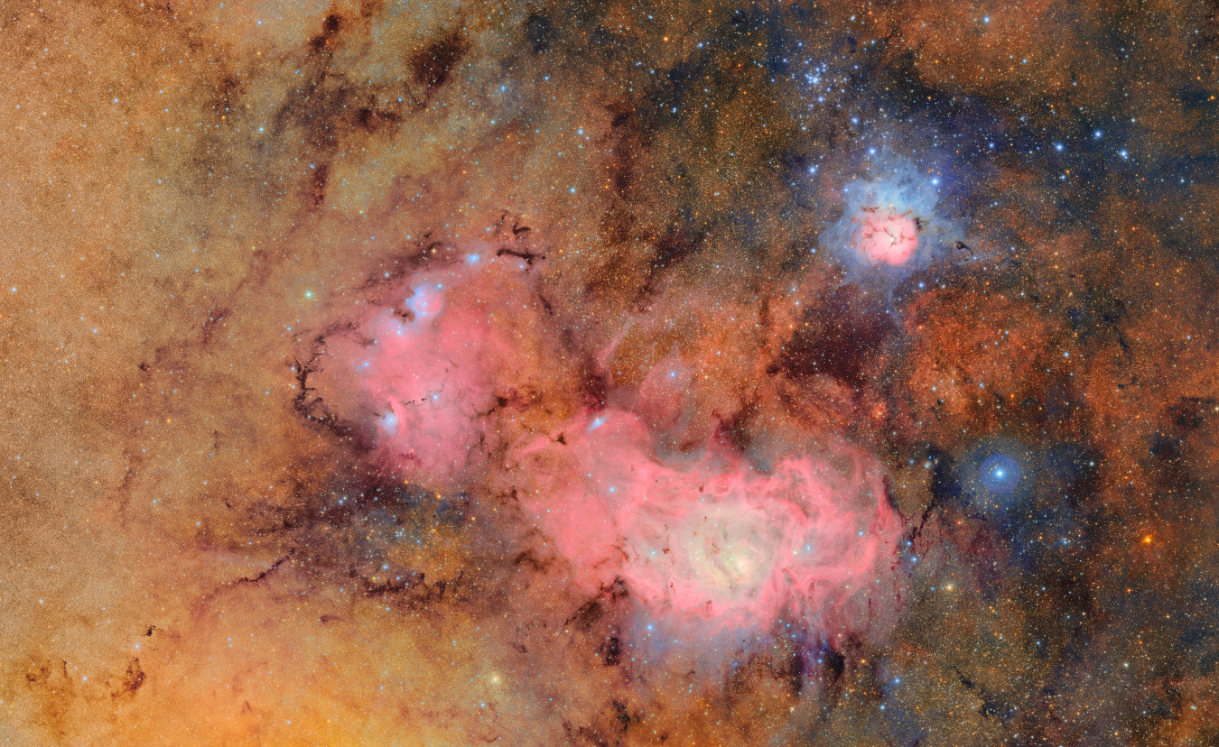 A tapestry of glowing tan and pink gas clouds with dark dust lanes. In the upper right, the Trifid Nebula resembles a flower with pinkish gas petals surrounded by blue gas, and streaked with dark, finger-like veins of dust. To the lower left, the much larger Lagoon Nebula stretches like a sea of magenta gas, with blue knotted clumps throughout. Both nebulae are embedded in a tan backdrop of gas that is brighter on the left than the right, etched with tendrils of dust and sprinkled with stars.