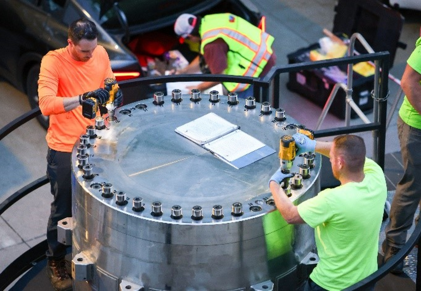 Two men word to turn two of the two dozen large bolts on a secure, metal container. Two other men work in the background. They all wear high-visibility clothes and vests.