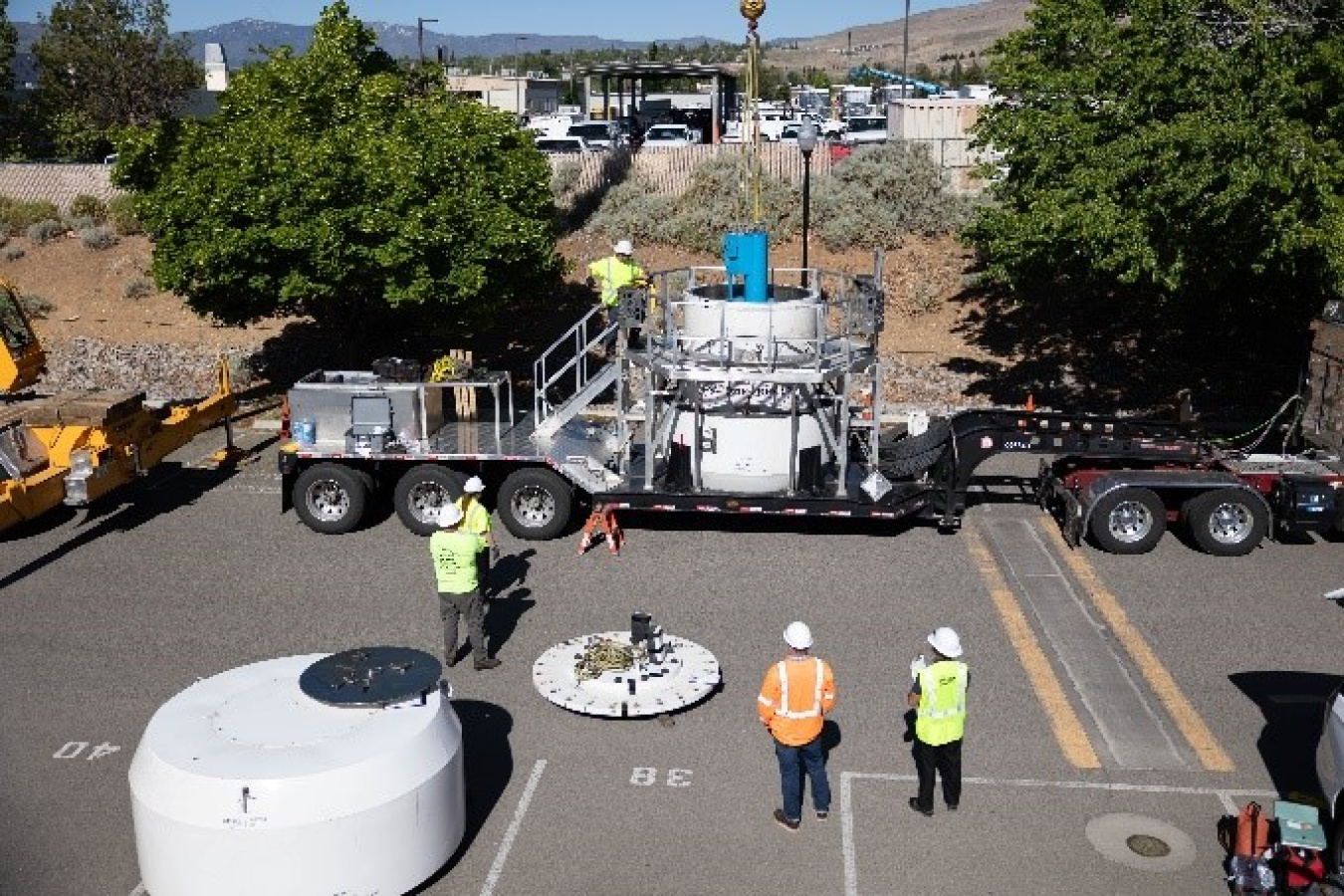 A photo of a blue irradiator being lowered by a crane into the transportation cask. It's taken from far away, so the entire trailer is visible. The trailer is sitting in a large parking lot. Four men, dressed in high-visibility clothing, watch.