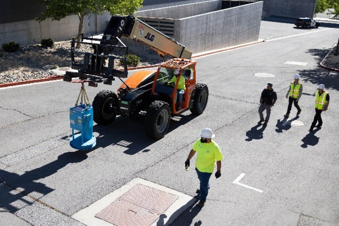 A small construction vehicle carries the irradiator in front of it as it travels into a parking lot. Four men dressed in high-visibility clothing look on.
