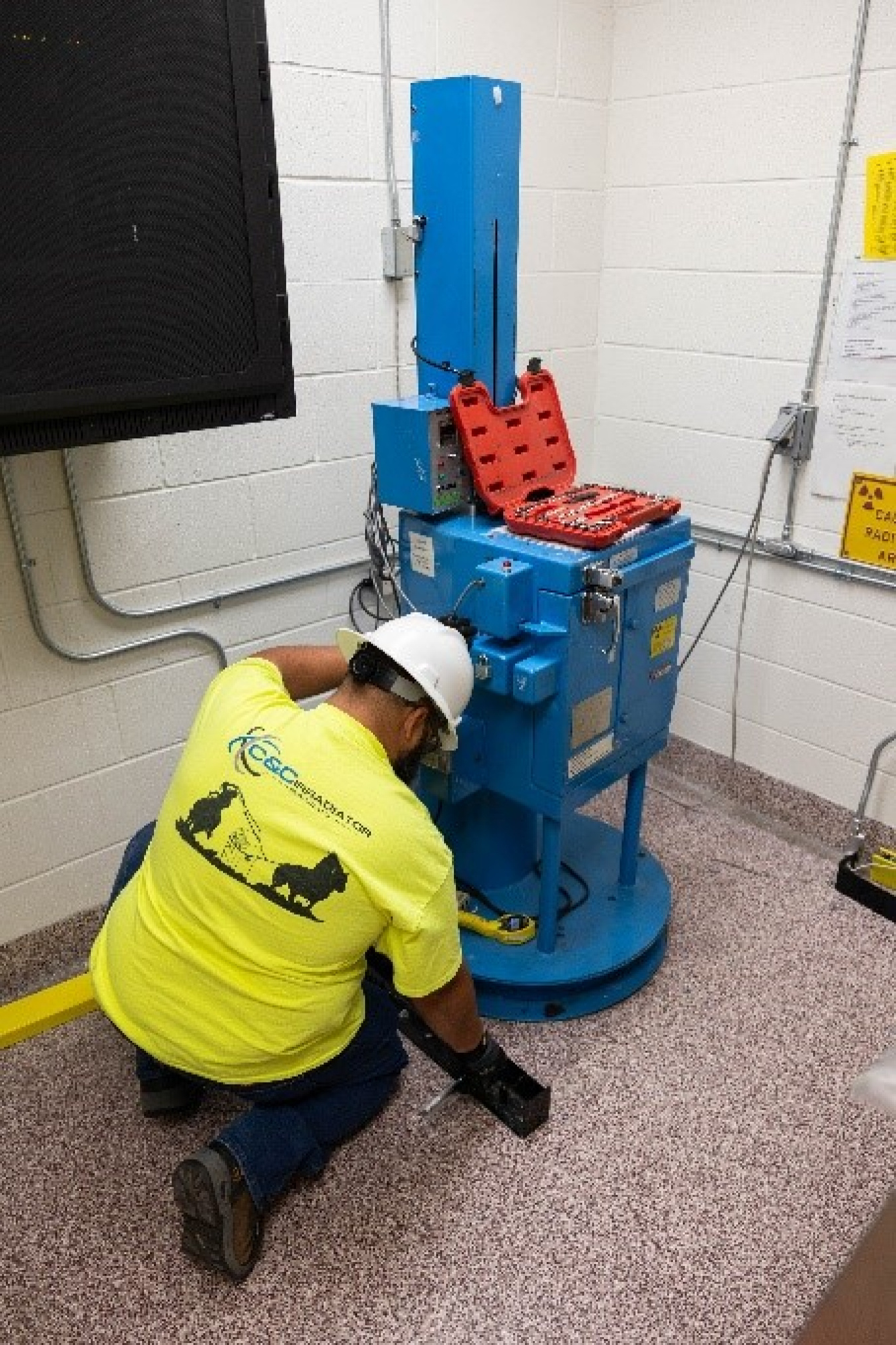 A man in high-visibility clothing kneels down on the floor. In front of him is a blue metal box about as big as a small refrigerator. He is working on it. His opened red toolbox sits atop the blue box.