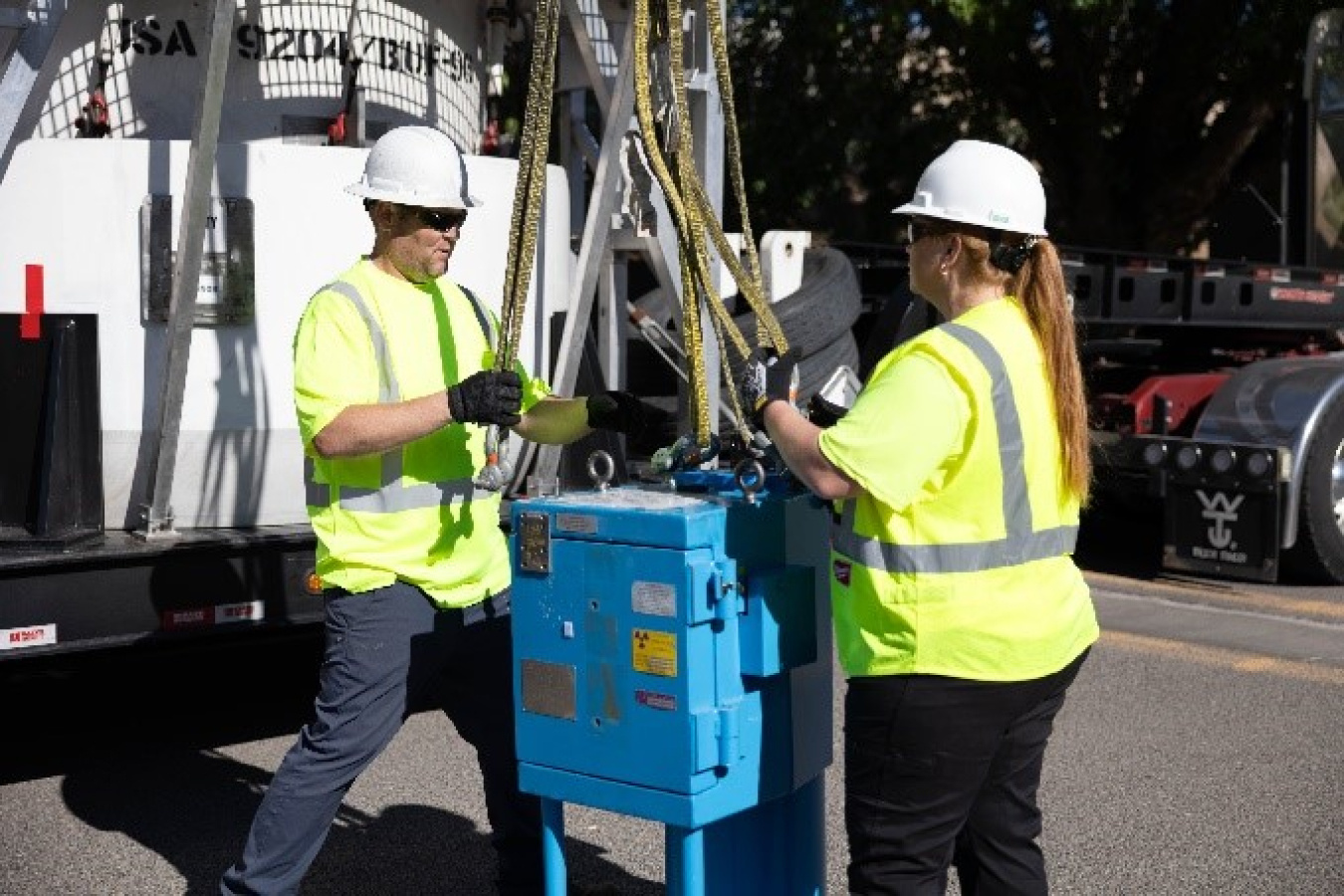 While it is hanging in the air, a man and a woman, both dressed in hard hats and high-visibility clothing, manipulate the straps at the top of the blue irradiator.