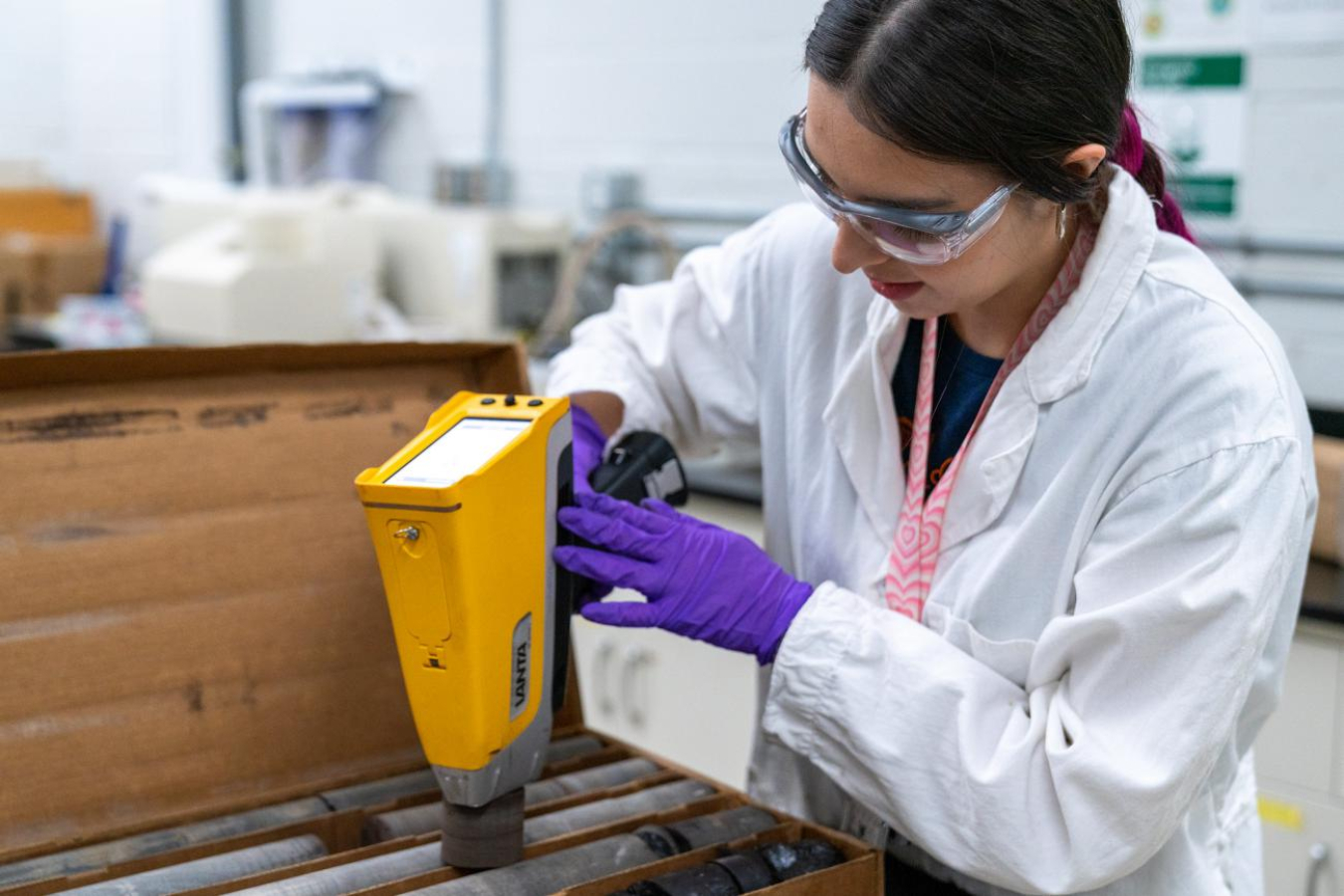 Worker in lab coat uses handheld sensing tool.