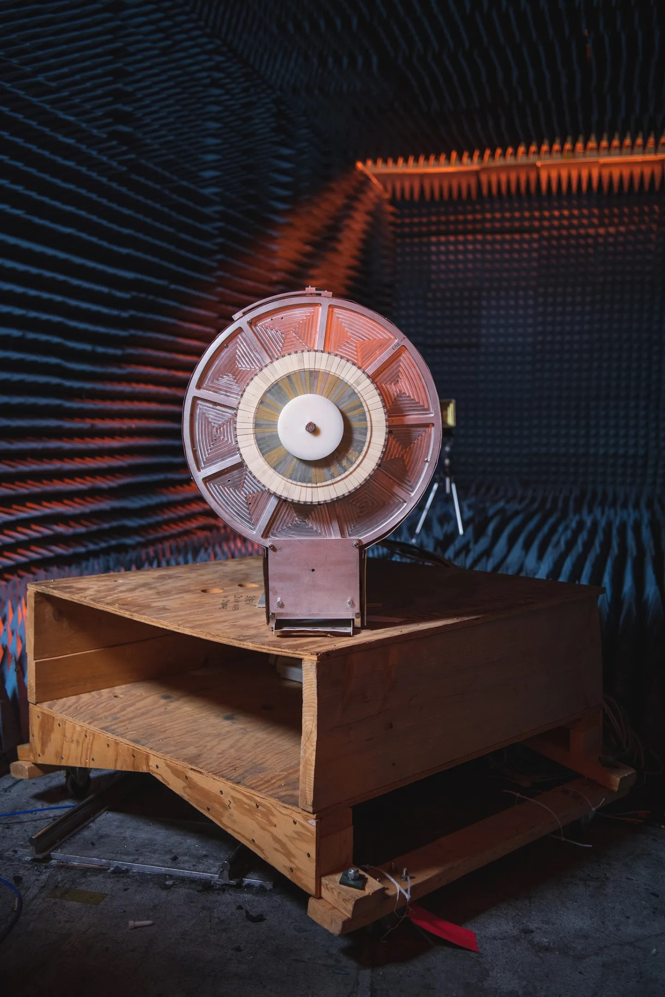 An object shaped like and about as big as an oscillating fan sits atop of wooden table. Its round part has equally shaped wedges that are ridged. This whole apparatus sits inside a so-called "quiet room."