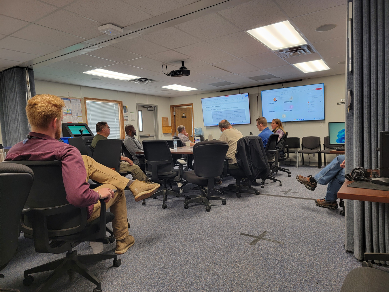 Employees in a conference sit at a chairs and observe screens at the front of the room