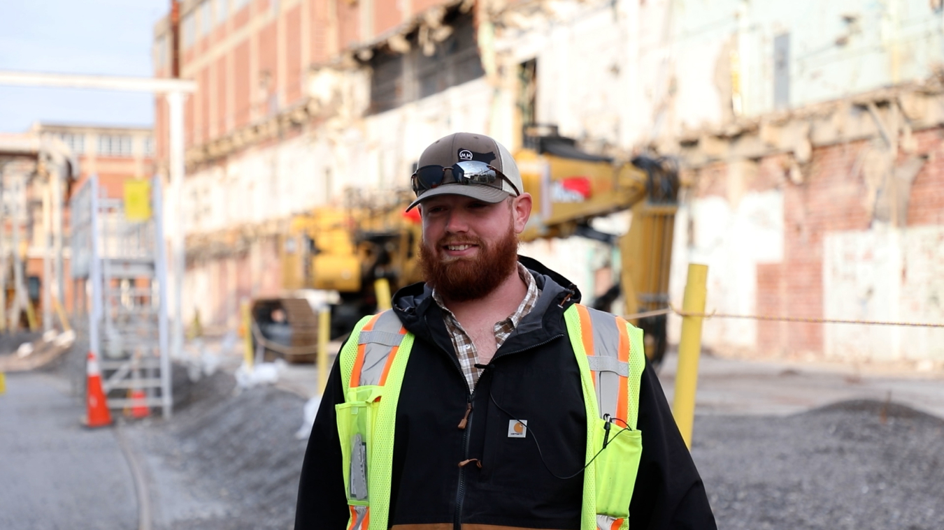 A man in protective gear smiles for a photo outside a demolished facility building