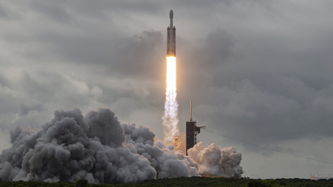 A rocket lifts off from a launch tower, with flaming thrust driving it higher. Smoke and steam billow below and to the left of it.