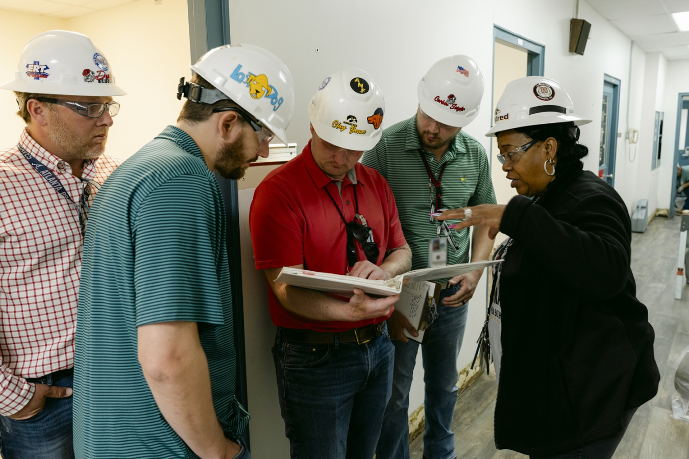 A group of employees in white hard hats look at a binder in a hallway