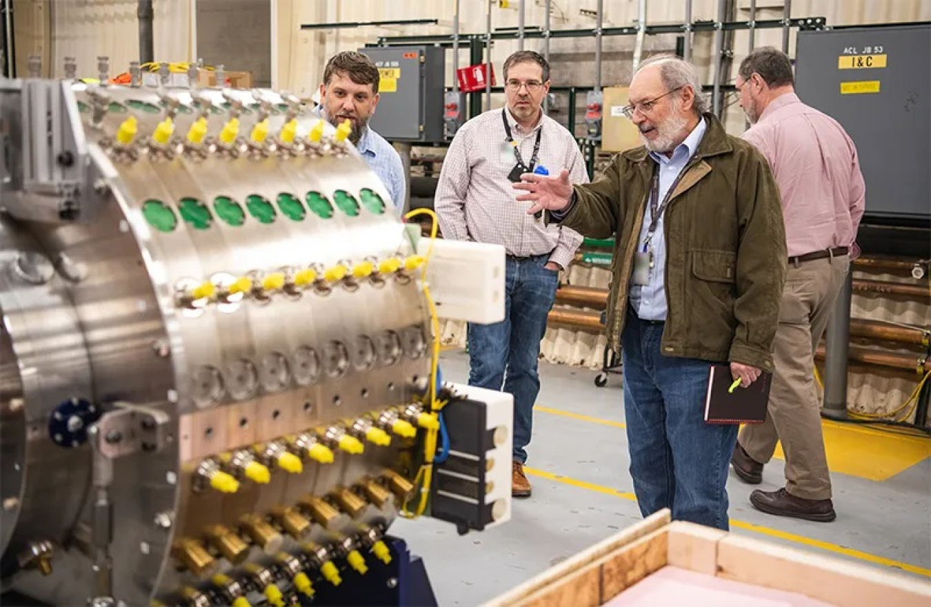 A man dressed in a jacket and holding a notebook gestures to a large metal mechanism that looks electrical. It has dozens of yellow bulb-like items sticking out in rows.