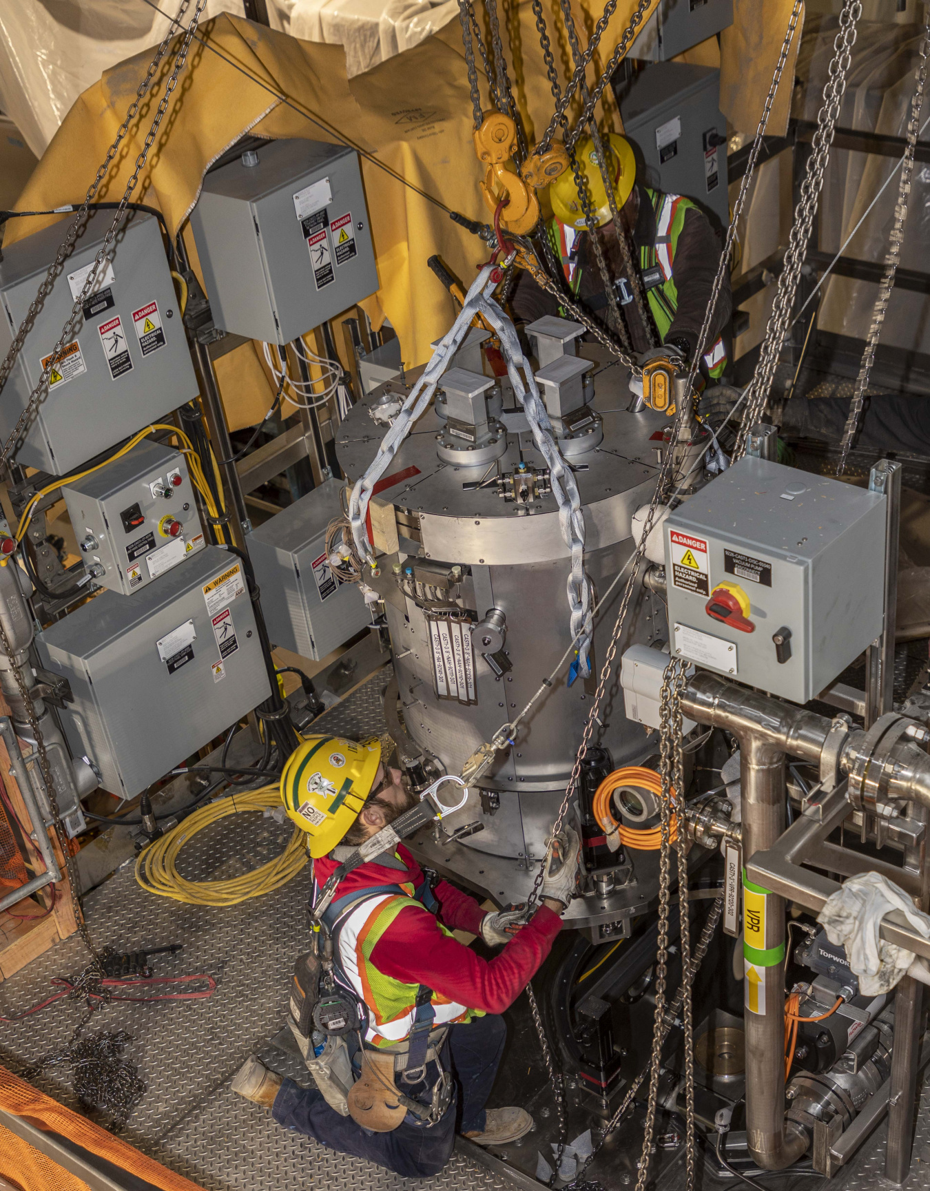 In a room crowded with electric and electronic equipment, a technician wearing a yellow hat, reflective vest and red shirt maneuvers a large metal apparatus into place. It is hanging on a chain, made of metal, and as large as an outdoor trash can.