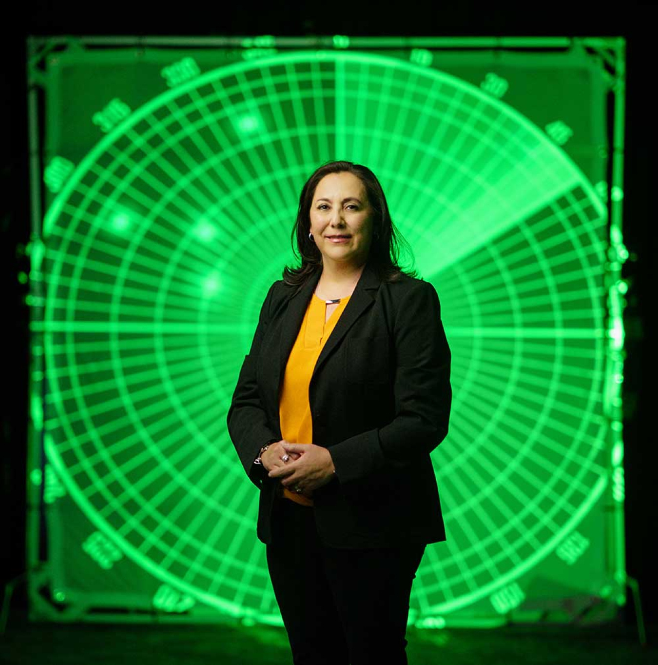 A woman dressed in black stands in front of a large, radar-looking green display