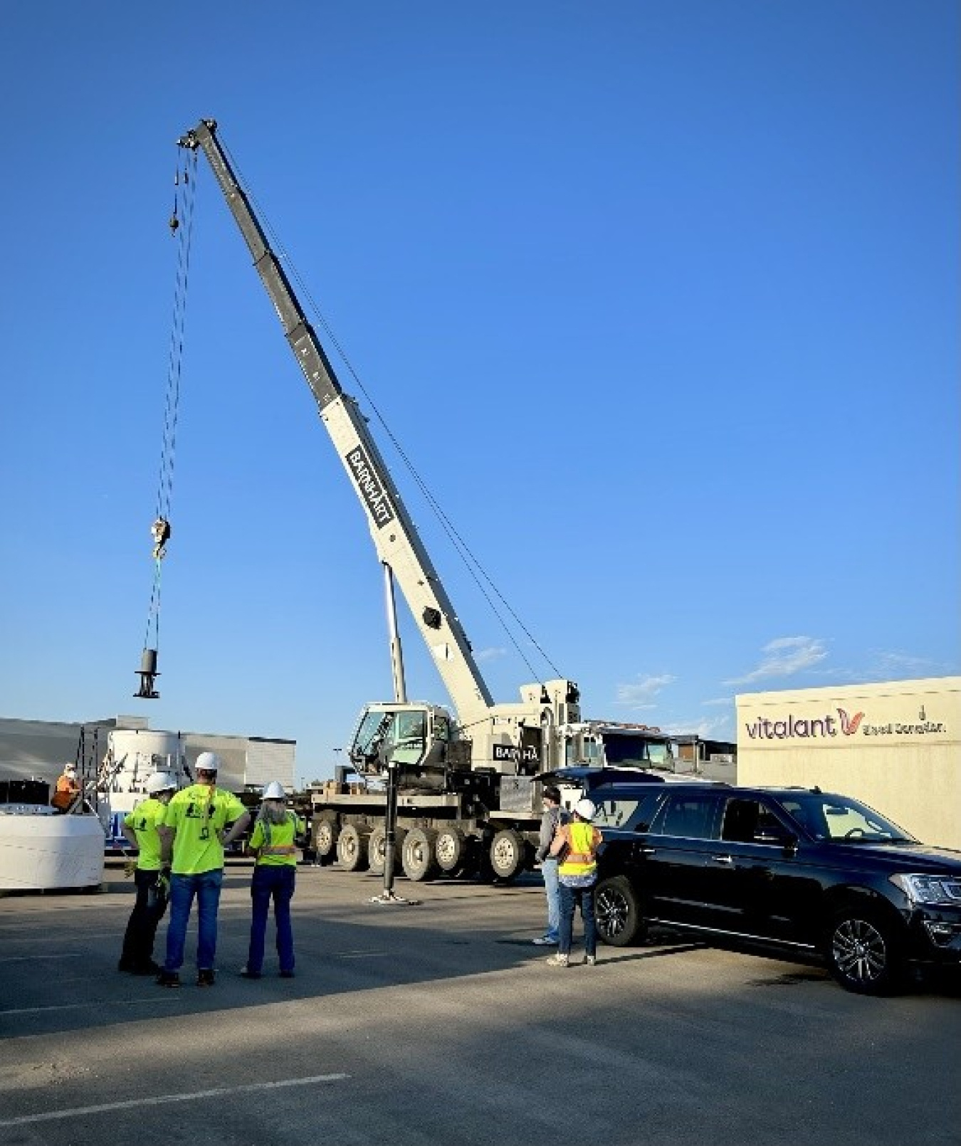 A large crane sets a container onto a flatbed tractor trailer. The crane is sitting in a parking lot and four people in high-visibility clothing watch it from a distance.