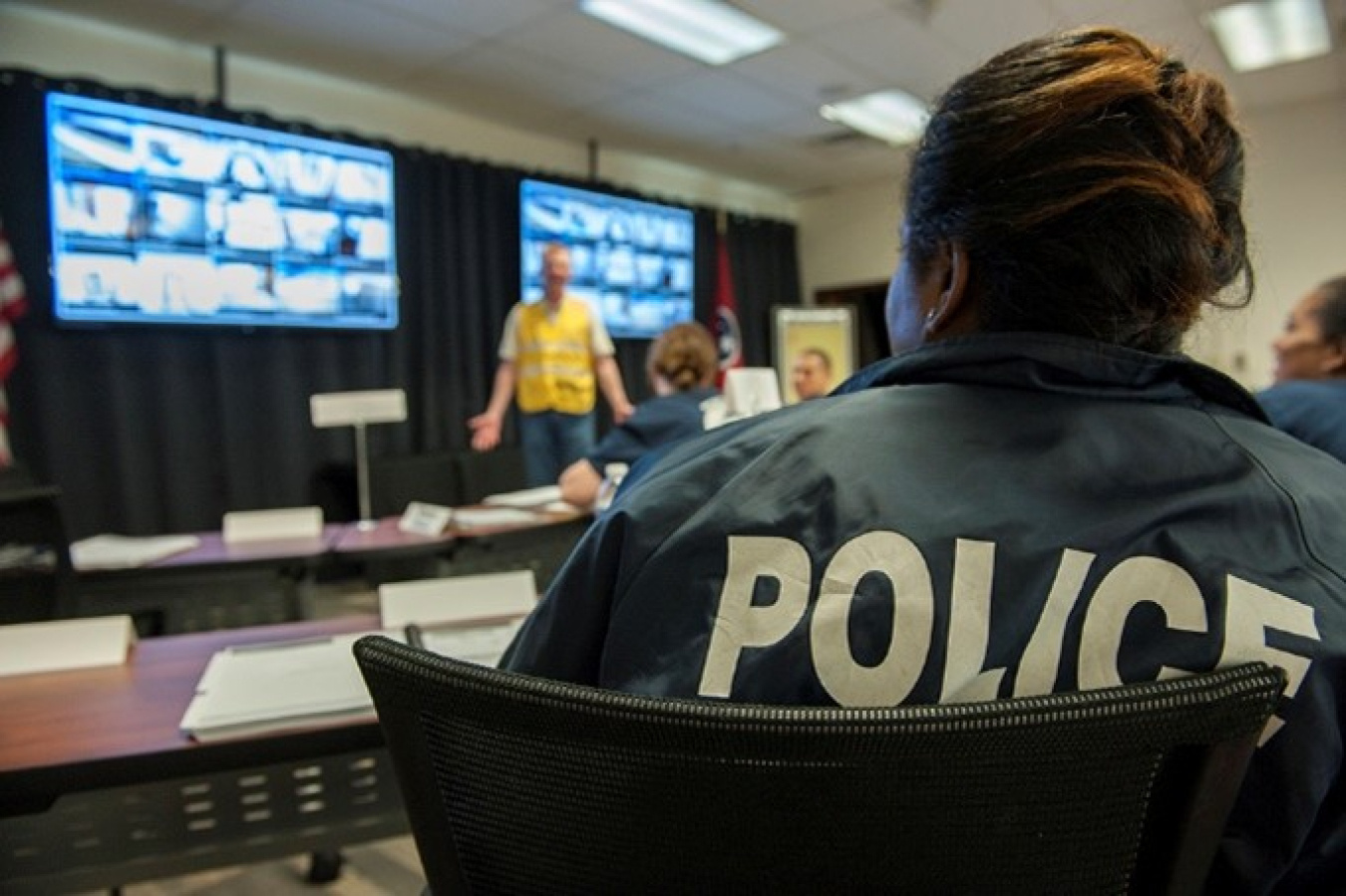 Shown from the back, a woman wearing a jacket emblazed POLICE watches two television screens while she is in the audience during a presentation. A presenter is at the front of the room.